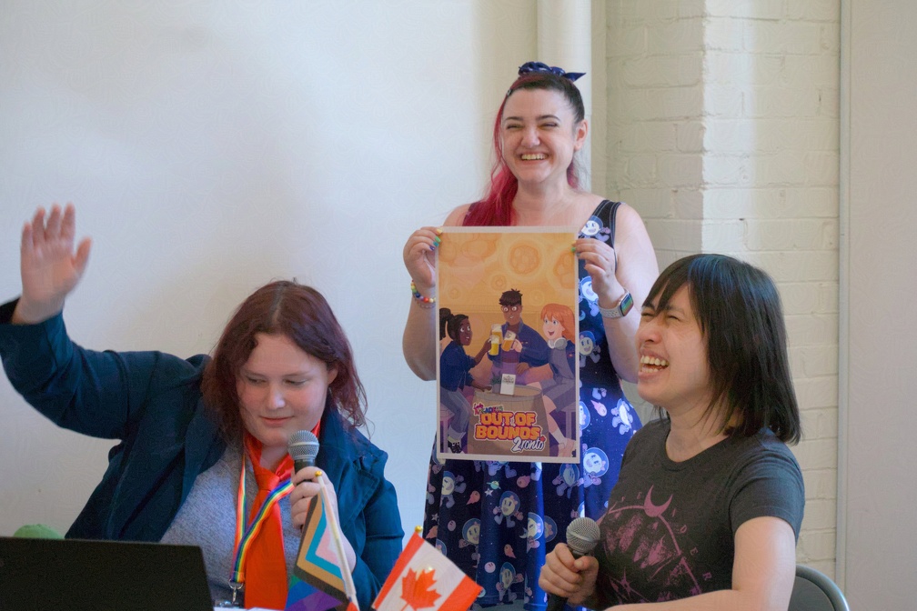 Two women sitting at a desk with a third standing behind them, holding up a poster at Lady Arcaders: Out of Bounds 2 2025. Photo by Jayde Iris Callejas. Two women sitting at a desk with a third standing behind them, holding up a poster.