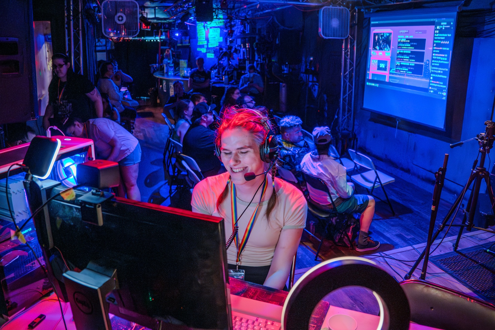 A smiling woman sits at a computer with an audience in the background watching a projector screen.